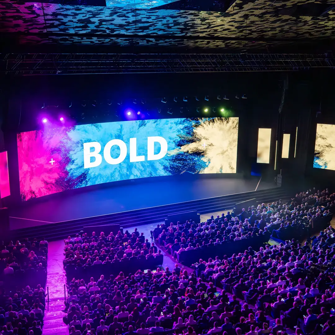 Large conference presentation with a bold visual slide displayed across multiple stage screens in front of a seated audience.