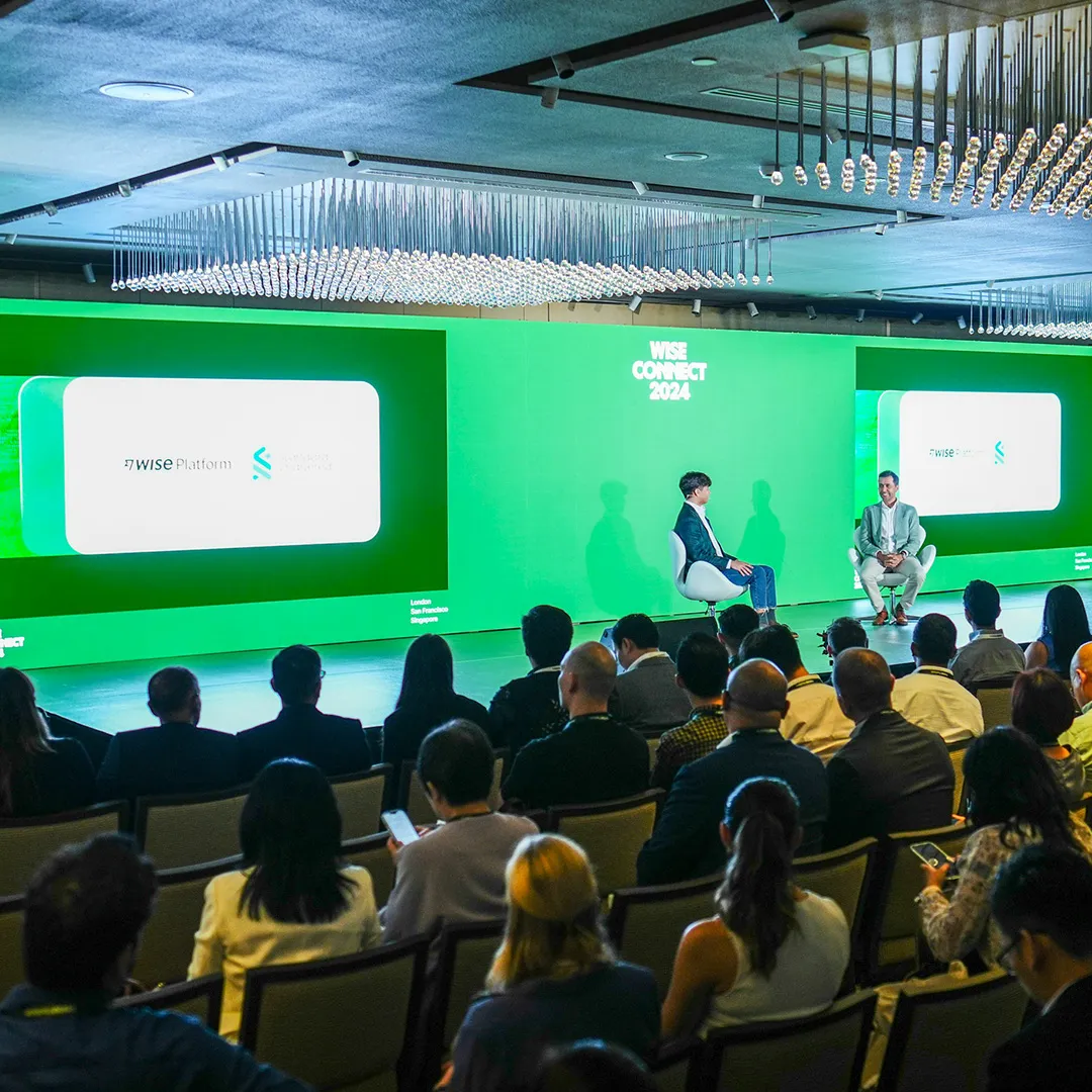 Audience watching a panel discussion on a bright green conference stage with large presentation screens.