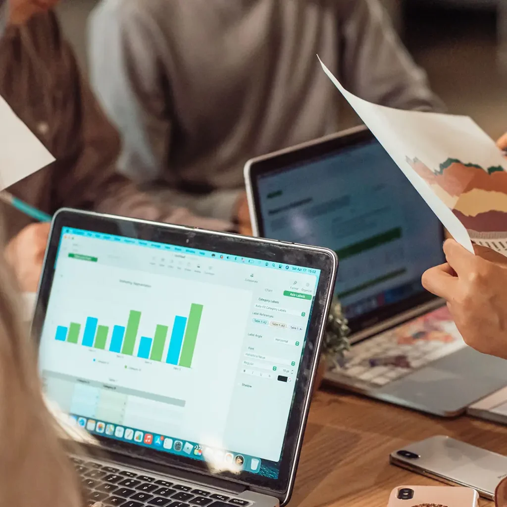 Team reviewing data visualisation charts on laptops and printed reports during a collaborative business meeting.